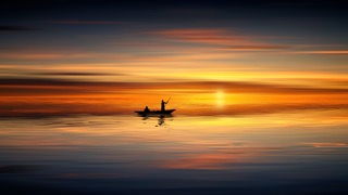 Two figures on a small boat silhouetted against an orange sunset reflected on smooth water. 